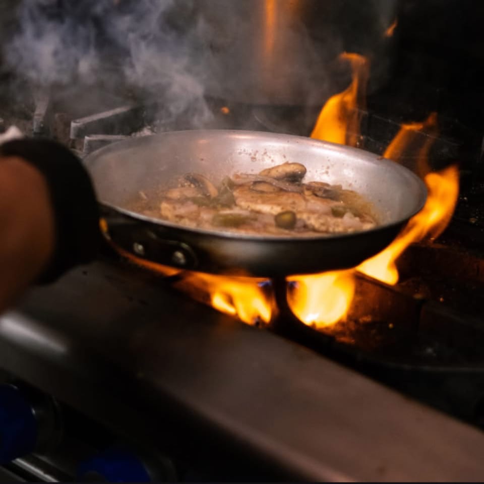 A chef's hand holds a pan of chicken and mushrooms cooking over a fiery stove.